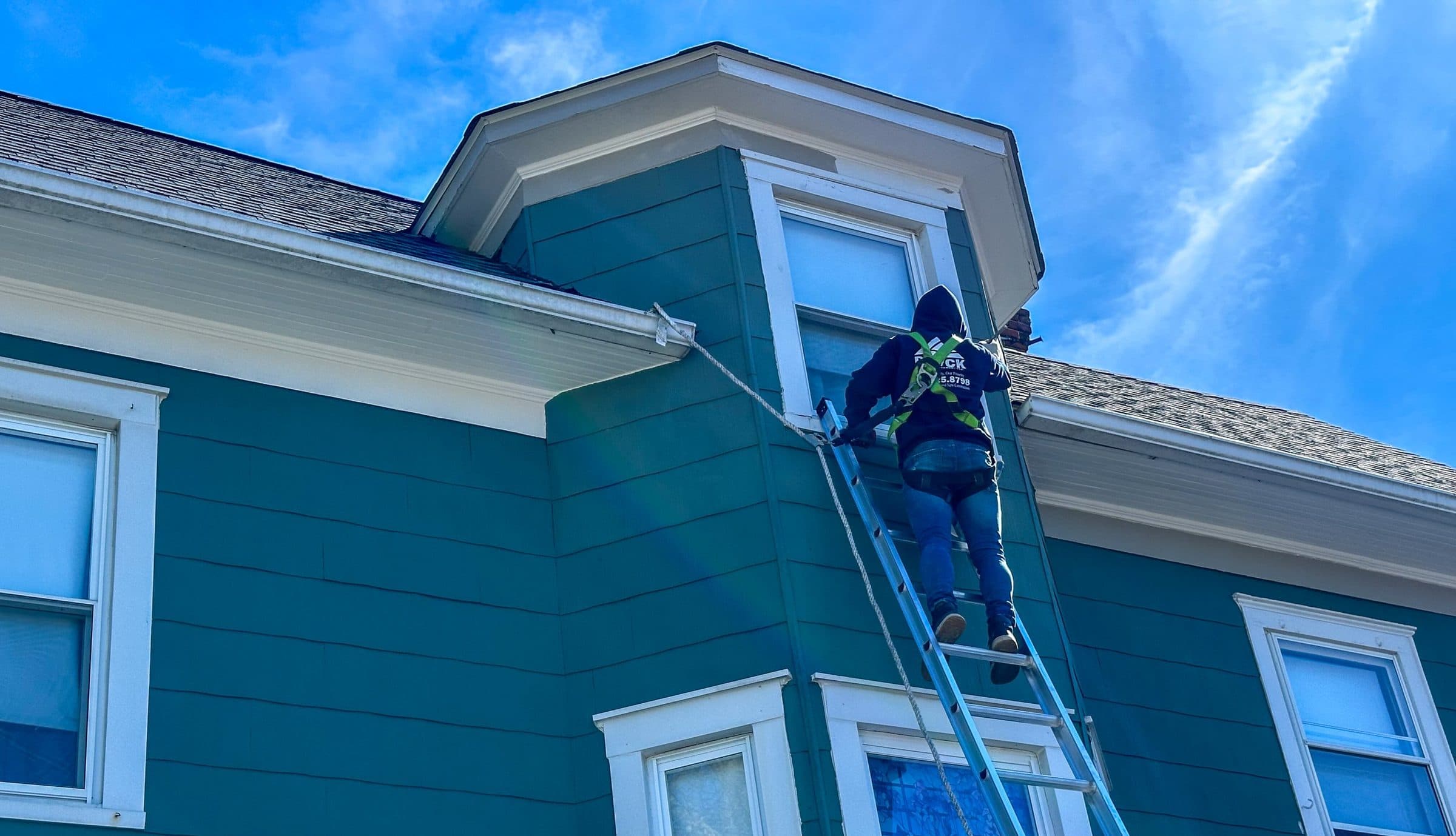 Crew member in a safety harness working on a ladder against a green-sided house